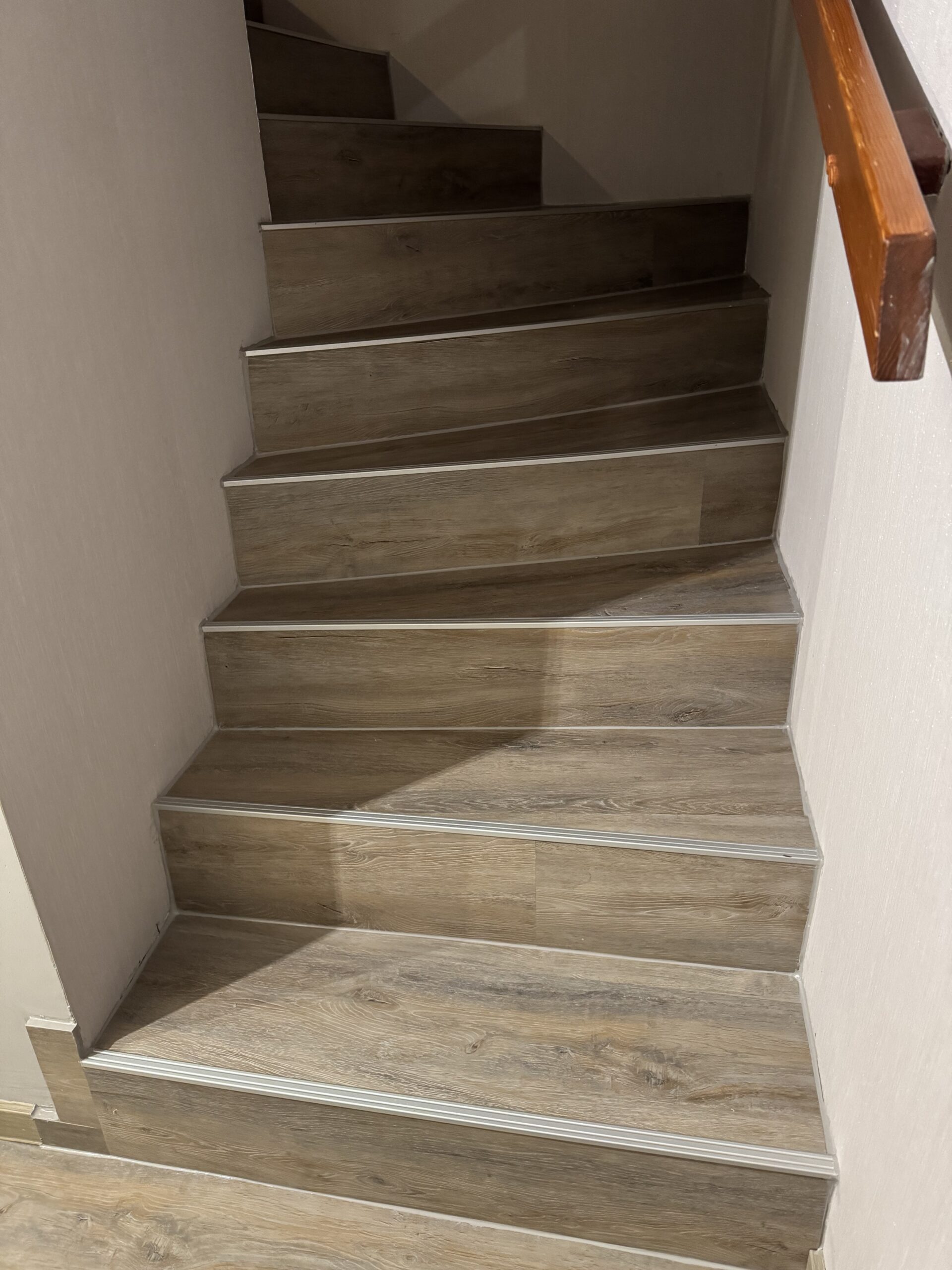 Gewendelte Treppe mit Vinyl in graubrauner Holzoptik, silberfarbenen Kantenprofilen und Holzhandlauf rechts